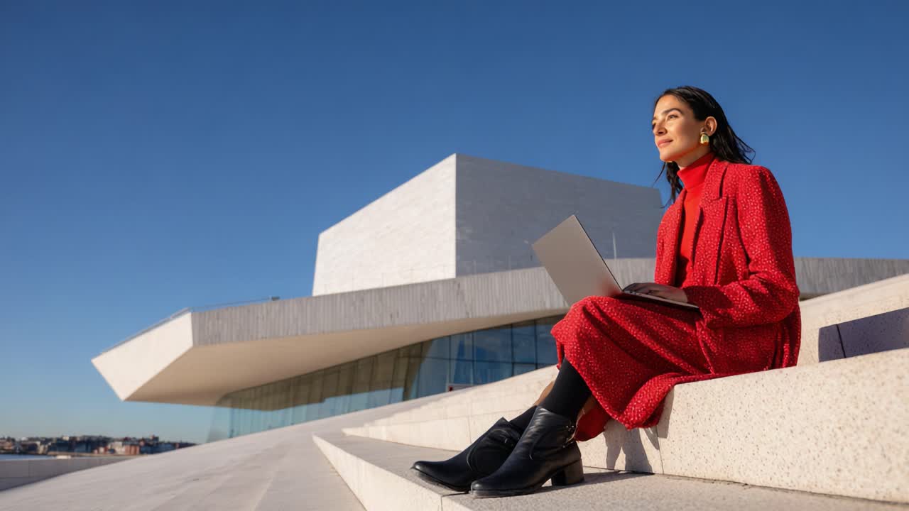 A professional woman in a stylish red outfit sits outside a modern building, engaged with her laptop while enjoying the clear sky and innovative architectural design