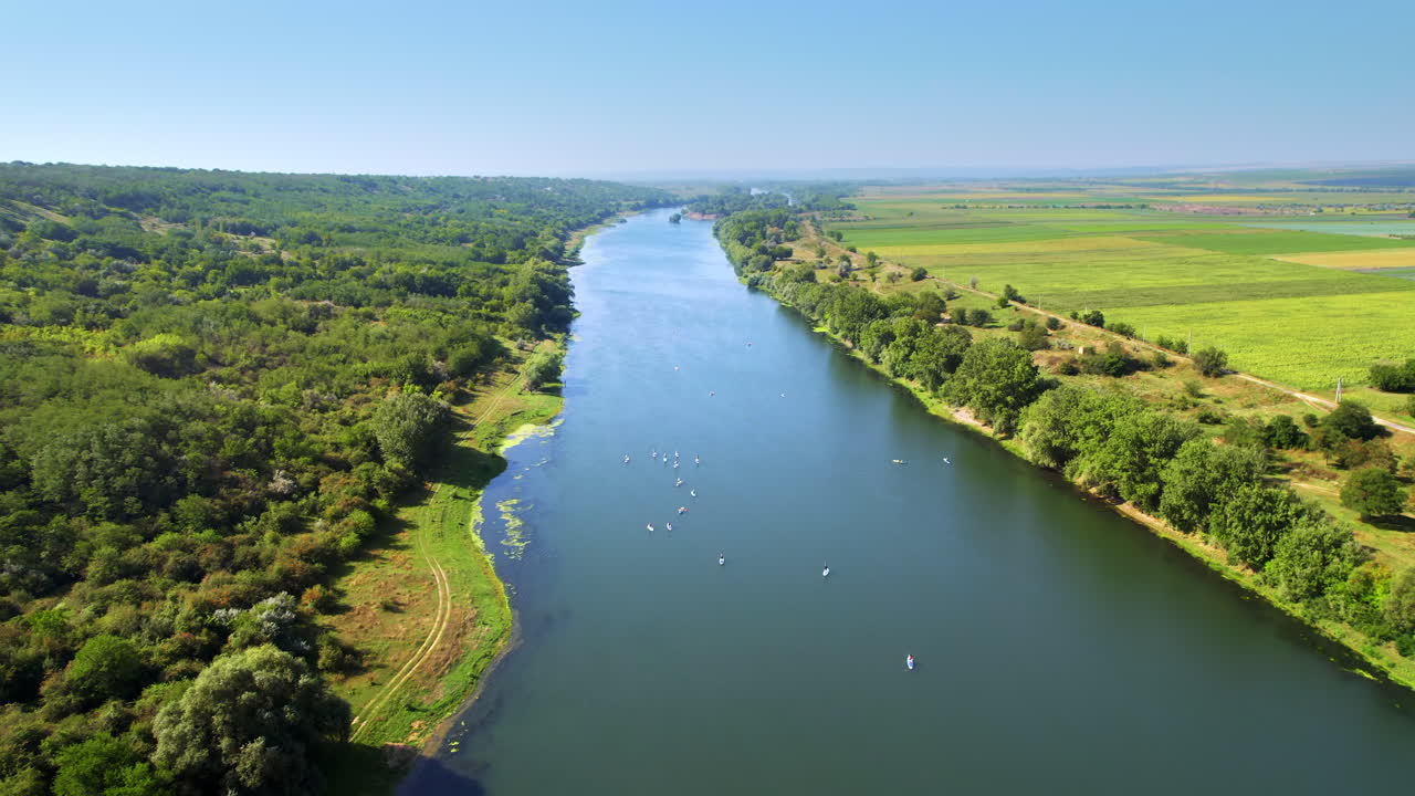 Letecký dronový pohled na více lidí, kteří dělají stand up paddleboarding na Dněstru v Moldavsku. Bujný les na obou stranách řeky, pole a vesnice