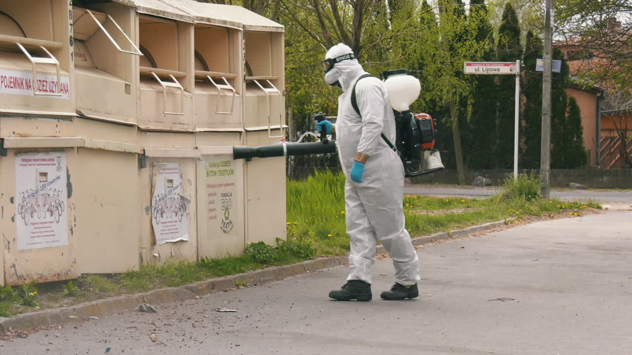 Disinfecting the garbage bins on public location. Man in protective suit using spray gun
