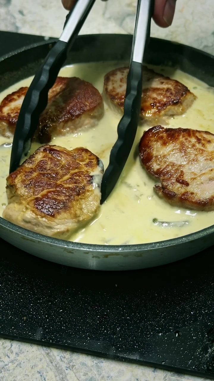 Close-up of pan-grilled pork tenderloin medallions simmering in a bubbling white peppercorn sauce. Vertical 4K cooking footage with rich texture and appetizing detail.