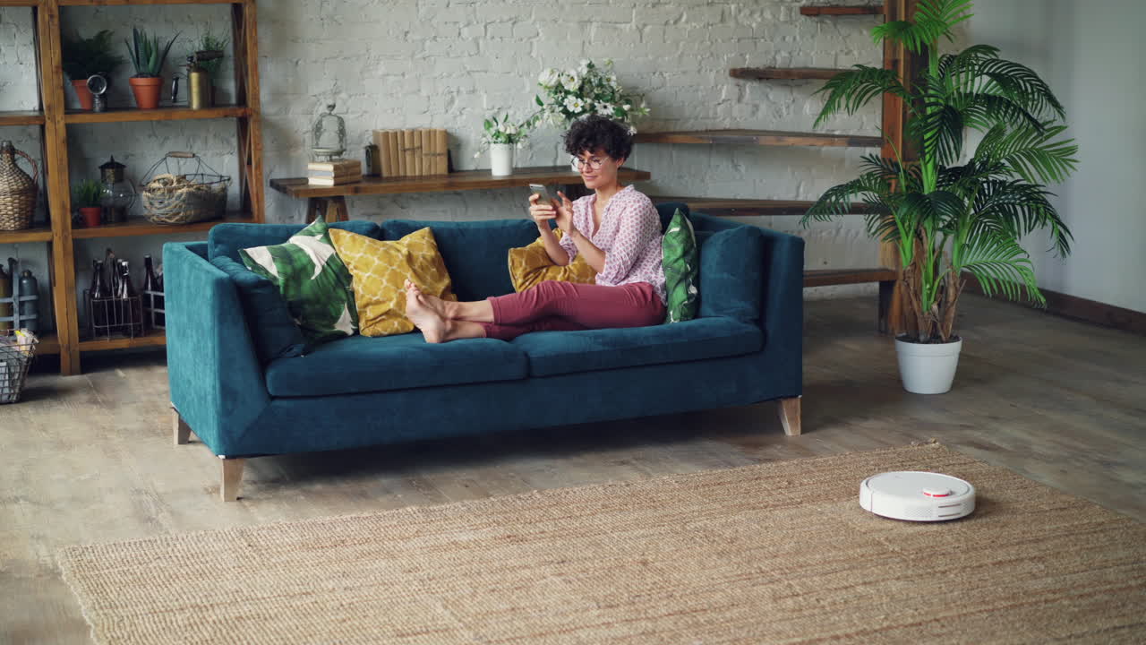 Woman Relaxing on Sofa Using Smartphone with Robot Vacuum