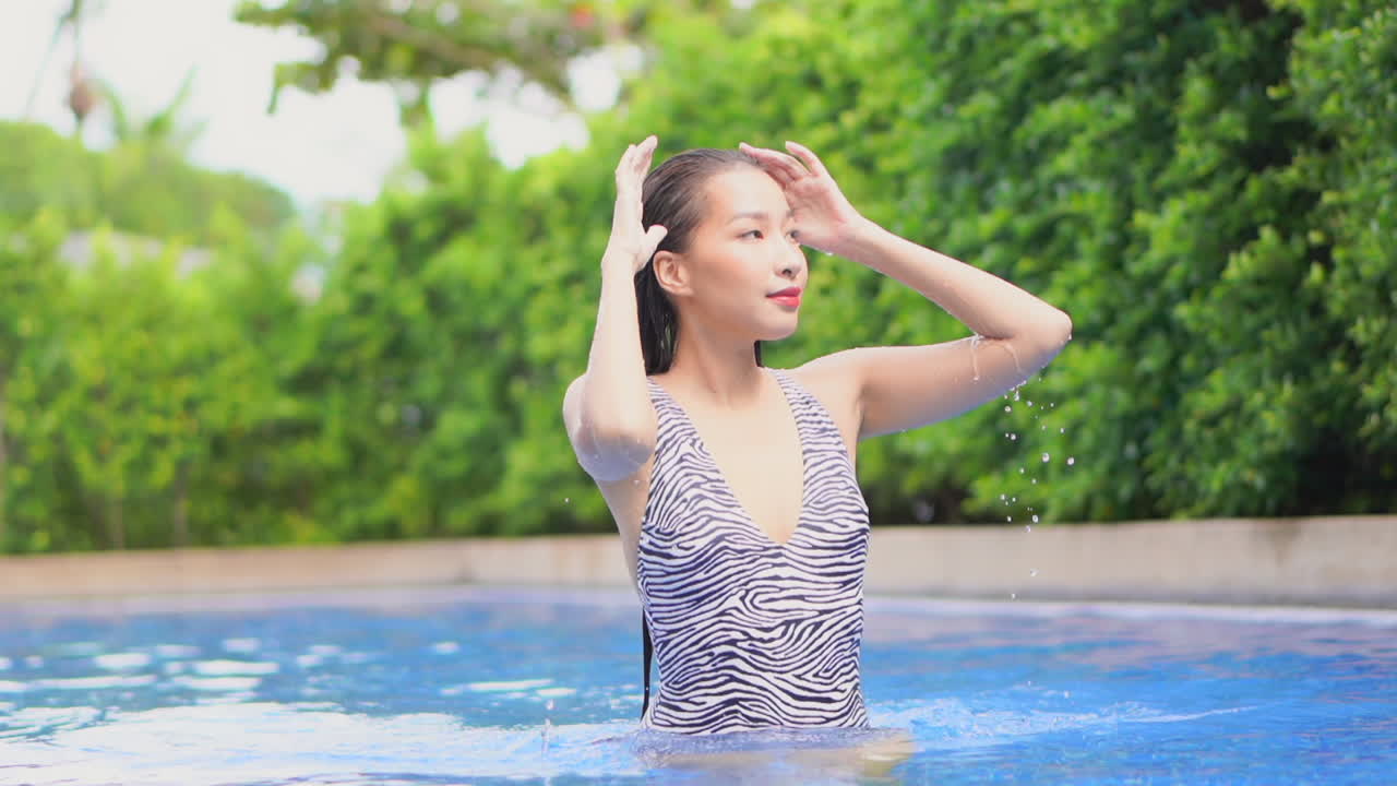 얼룩말 무늬 수영복을 입은 예쁜 젊은 여성이 수영장 물을 통해 카메라를 향해 걸어가면서 젖은 머리를 부드럽게 다듬습니다.