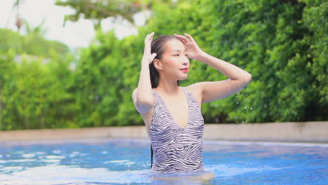 a pretty young woman in a zebra print swimsuit smooths down her wet hair as she walks through the pool water toward the camera