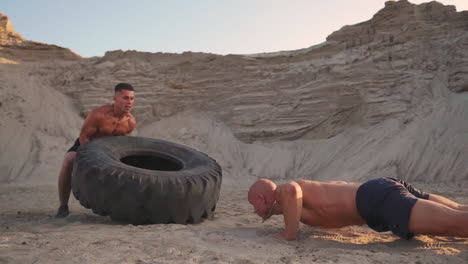 two muscular open-chested athletes train in active mode on the beach doing push-ups and pushing a huge wheel against a sandy mountain at sunset