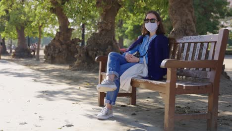 mujer joven sentada en un banco del parque con una máscara protectora durante la pandemia de covid 19