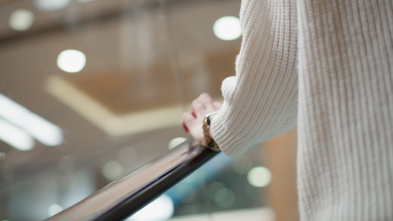 close up of demure woman wearing wrist watch tapping handle of moving walkway in modern mall corridor under bright overhead lights beside glass railing capturing gentle motion and casual elegance