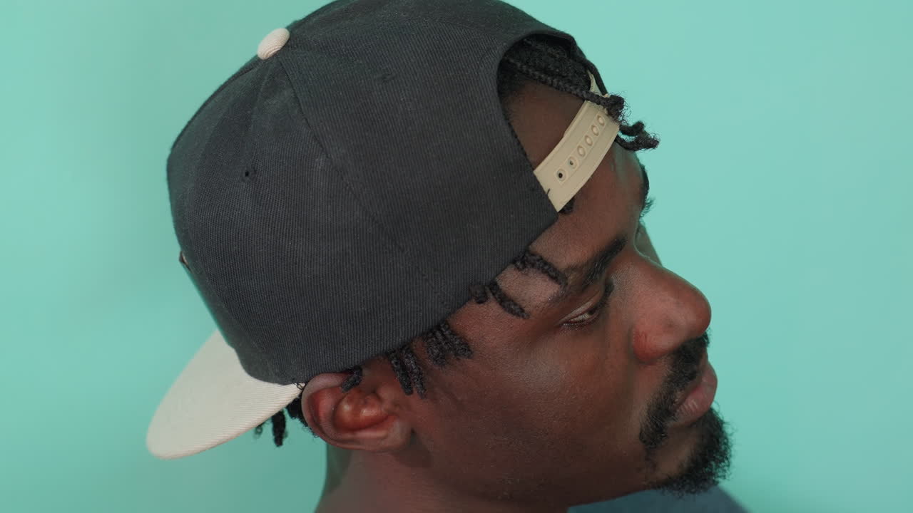 Close up side profile of dark skin man wearing black backward cap looking up thoughtfully with calm expression against mint green background
