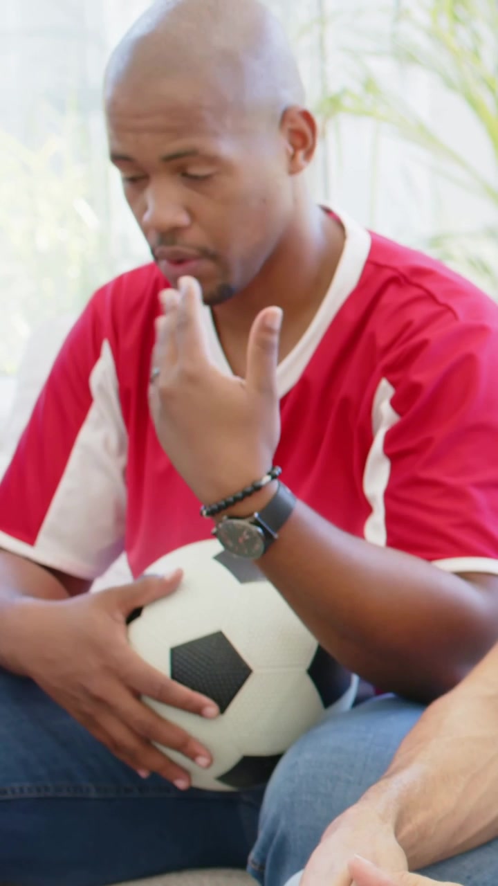 Video verticale: Telecamera panoramica che mostra un gruppo che discute di una partita sul divano mentre un uomo con una maglia rossa tiene in mano una palla.