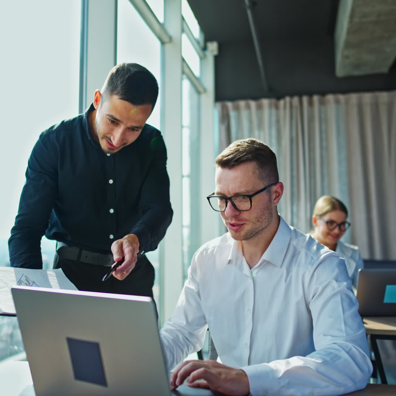 Male colleagues discussing work issues looking at papers. Female employee sitting at the desk behind. City panorama in the windows