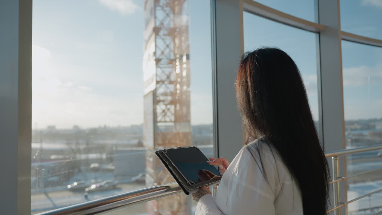 Back view of woman standing by glass window using tablet and slowly gazing up with calm expression as warm daylight fills modern indoor space overlooking city structures in soft blur
