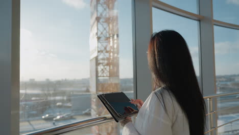 back view of woman standing by glass window using tablet and slowly gazing up with calm expression as warm daylight fills modern indoor space overlooking city structures in soft blur
