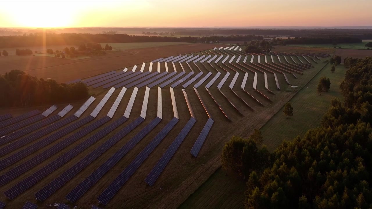 Droneoptagelser fra luften af en stor solcellepark nær Sigulda, Letland, ved solopgang, med rækker af fotovoltaiske paneler, der reflekterer varmt gyldent sollys hen over det landlige landskab