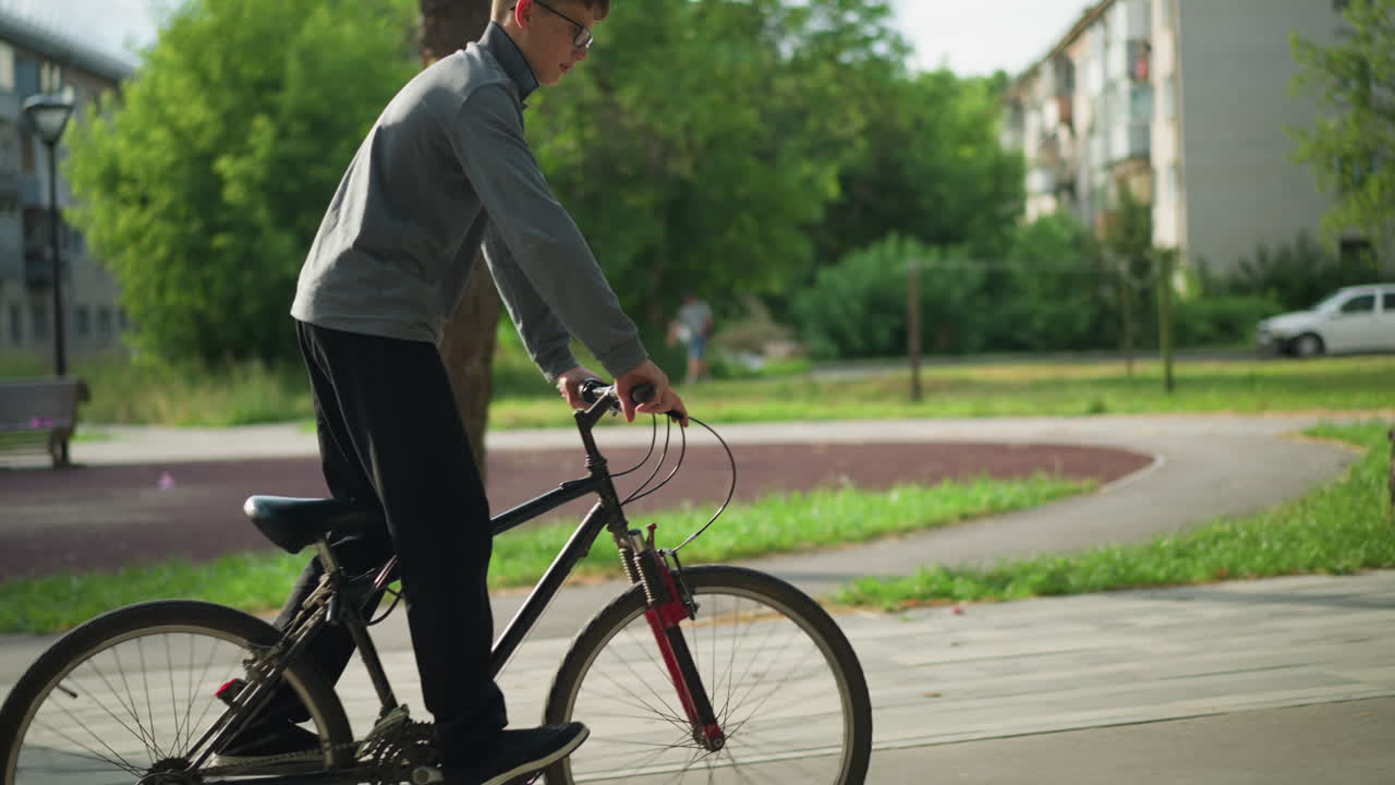 회색 과 검은색 바지를 입은 어린 소년은 느린 동작으로 공원에서 자전거를 타고 있습니다. 배경에는 흐릿한 건물, 나무, 주차된 자동차, 그리고 멀리서 고 있는 누군가가 있습니다.
