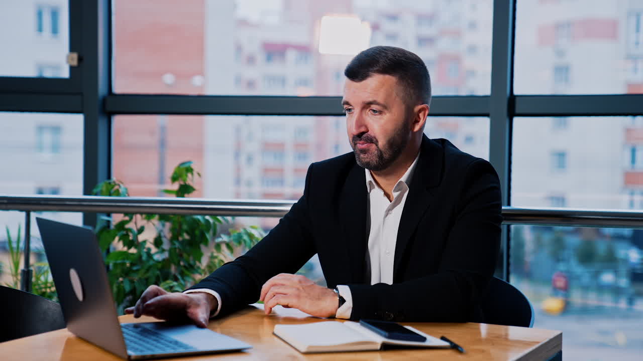 Businessman in suit working at his laptop. Man presses keys on keyboard and comments. Buildings at backdrop in blur.