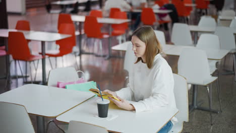 woman sitting at table in mall, reading her book with coffee cup on table and shopping bag close to her, soft, lively mall atmosphere with light blinking on the floor