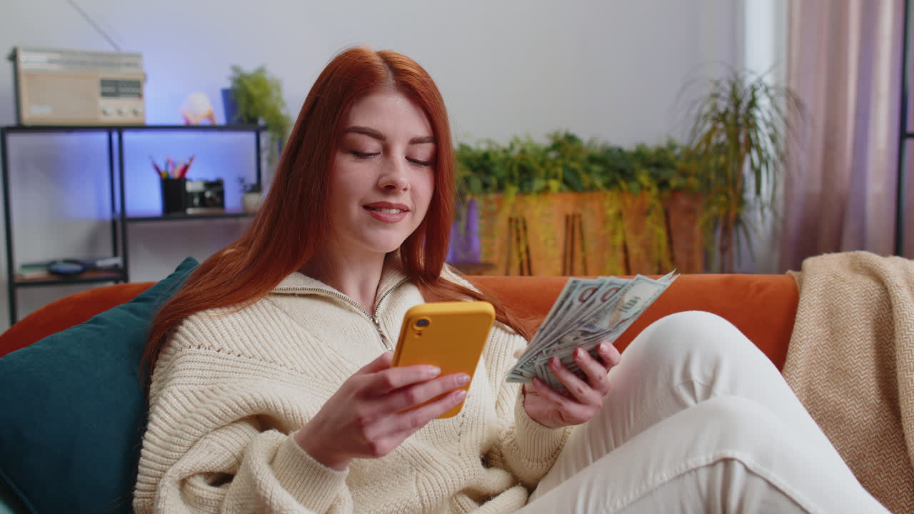 Smiling happy young woman counting money cash use smartphone income saves lottery win budget at home