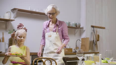 little girl helping her grandmother to set table for dinner and bringing a tray with fresh food 1