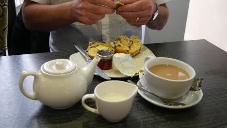 a man buttering a scone with butter and jam at a table set with a teapot, milk jug, and a cup of tea