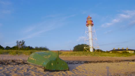 Beautiful-view-of-the-Pape-Lighthouse-and-green-coastal-fisherman-boat-on-a-calm-summer-evening-with-slow-moving-clouds-in-the-background-before-the-sunset,-wide-shot