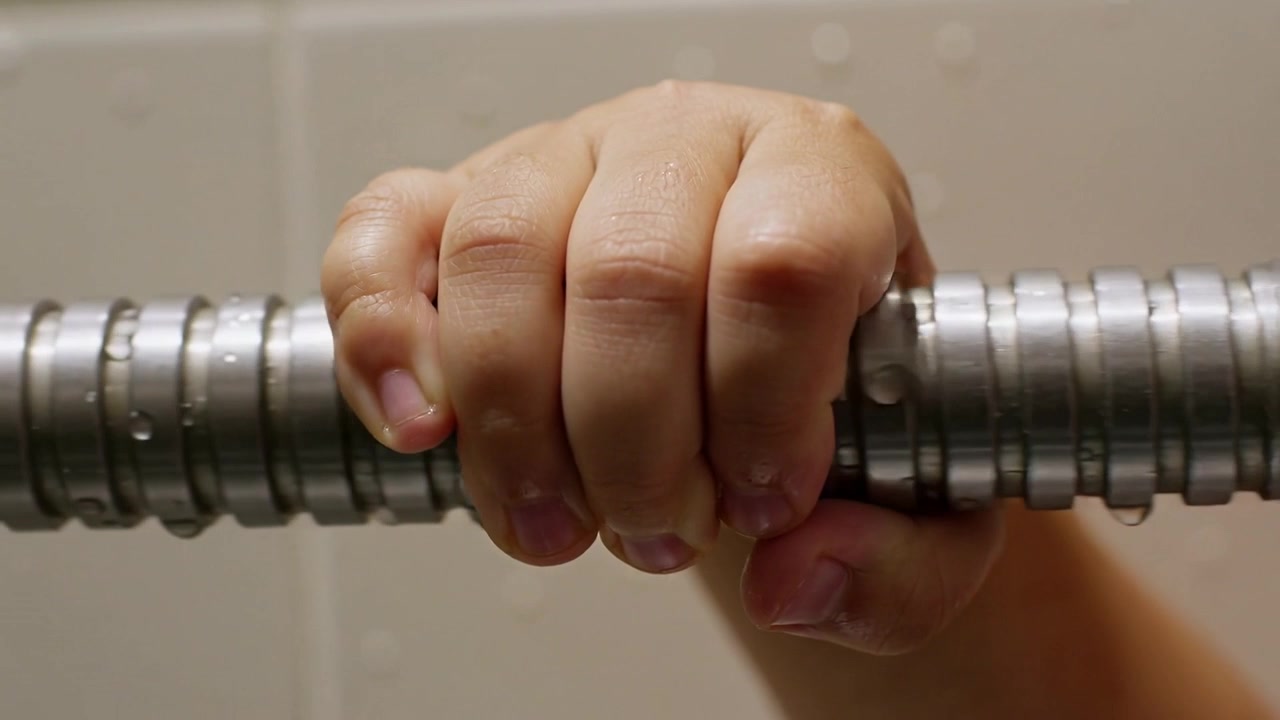 Mano di bambino che afferra saldamente un tubo doccia in metallo ondulato su una parete piastrellata mentre le goccioline scivolano.