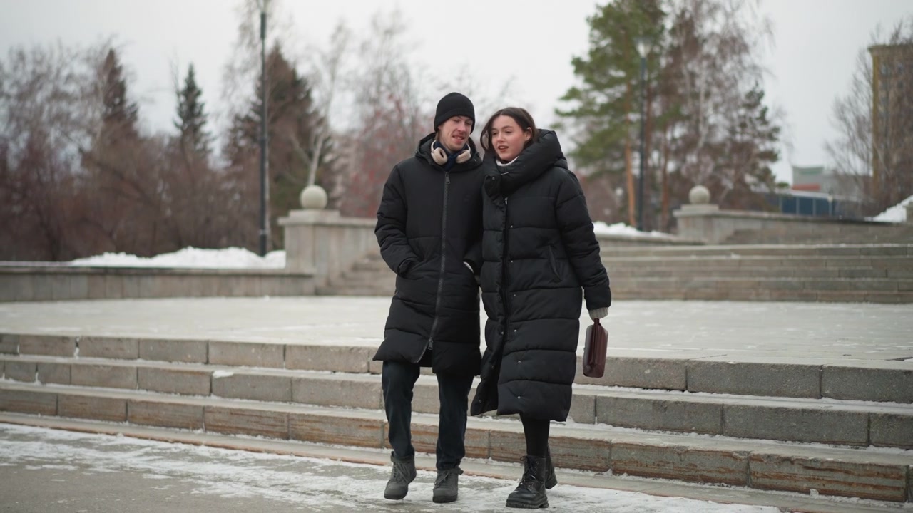 Two people walk together outdoors in winter wearing long black coats and boots. One carries small handbag while other has headphones around their neck. scene features snow covered ground