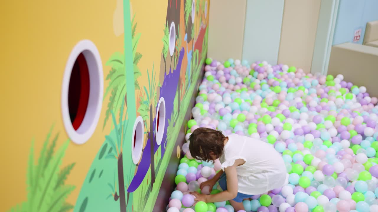 Cute little girl has fun throwing colorful plastic balls into target holes on a wall while playing in a large ball pit at an indoor playroom