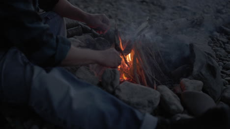 Medium-view-of-man-by-fire-pit-placing-small-sticks-around-tipi-campfire,-slow-motion-smoke-rises