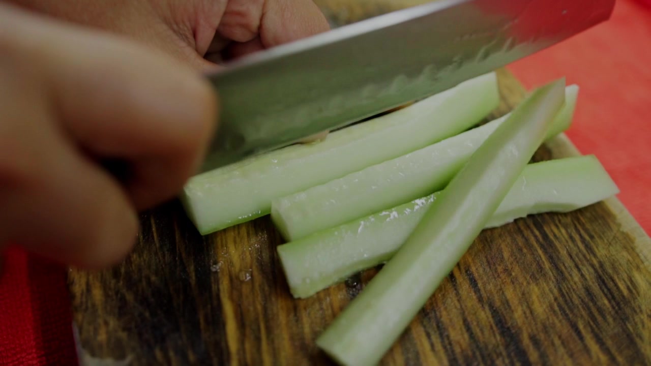 Closeup view of hands slicing fresh cucumber with kitchen knife on wooden cutting board, everyday food preparation scene showing healthy ingredient cutting for homemade cooking