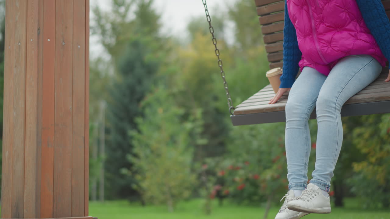 Donna bianca su una panchina a dondolo di legno in un parco ricco di vegetazione, sorseggia un caffè da asporto, indossa un gilet rosa e jeans di denim, penzola sneakers bianche, il bicchiere da asporto è appoggiato accanto, primo piano delle gambe e delle scarpe, le catene cigolano