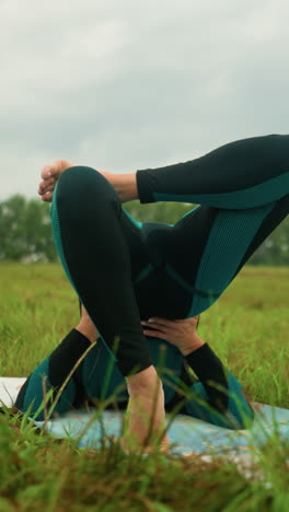 vista trasera de una mujer blanca acostada en una alfombra de yoga practicando la postura del puente con las manos apoyando su cintura en un campo cubierto de hierba bajo un cielo nublado, los árboles están alineados en la distancia
