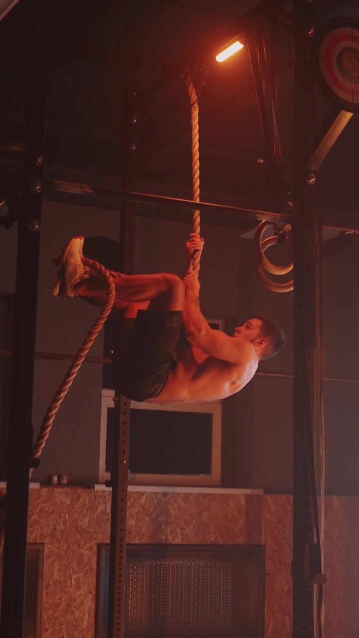 Uomo muscoloso e intenso che si arrampica alla corda in una palestra buia