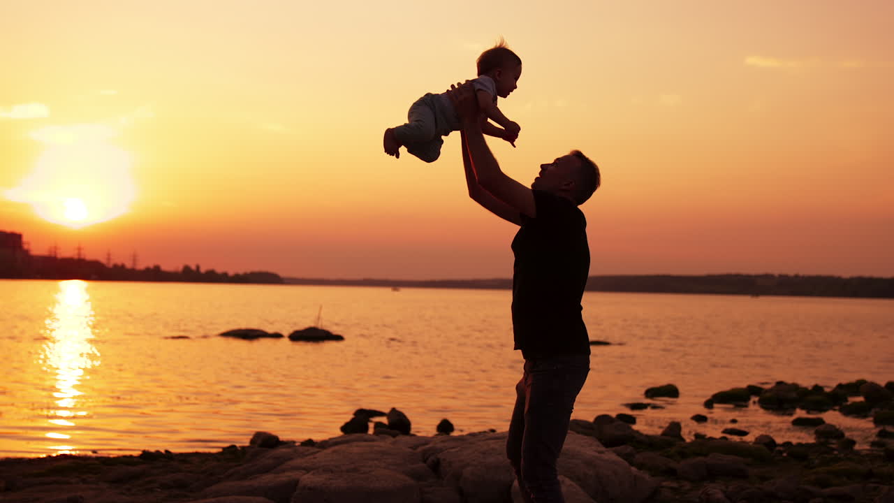 Careful dad playing with his baby at the river waterfront. Loving father tossing up his son and kid rejoices. Setting sun over water at backdrop.