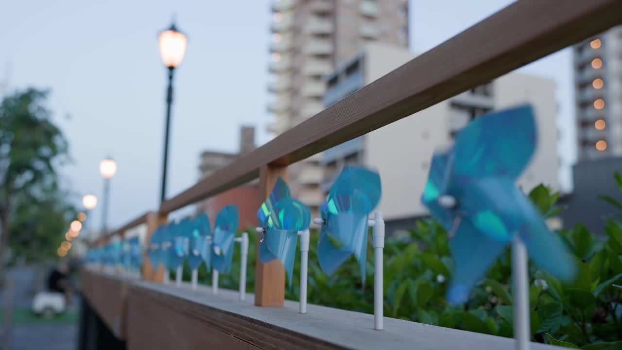 A close-up shot capturing blue pinwheels decorating the railing of the promenade at Tokyo Mizumachi along the Kita-jūkken-gawa River. The shot shows the pinwheels spinning in the wind