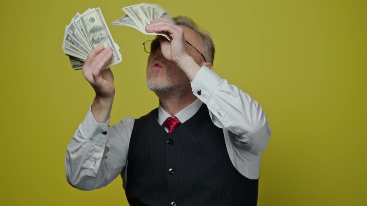Senior man with money in his hands. Happy rich man is waving dollar banknotes on yellow studio background. Middle aged man enjoys big amount of money.