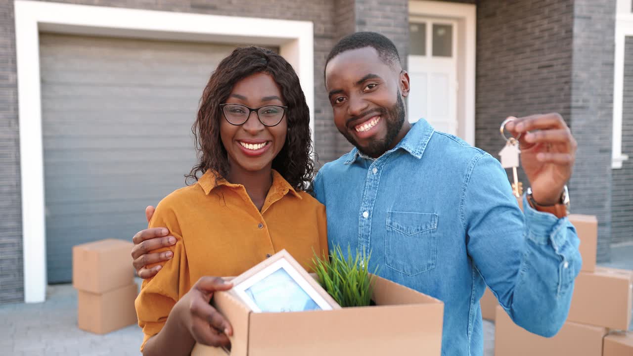 Portrait of African American young married couple of man and woman standing at new home, holding carton box with stuff and showing key when moving in