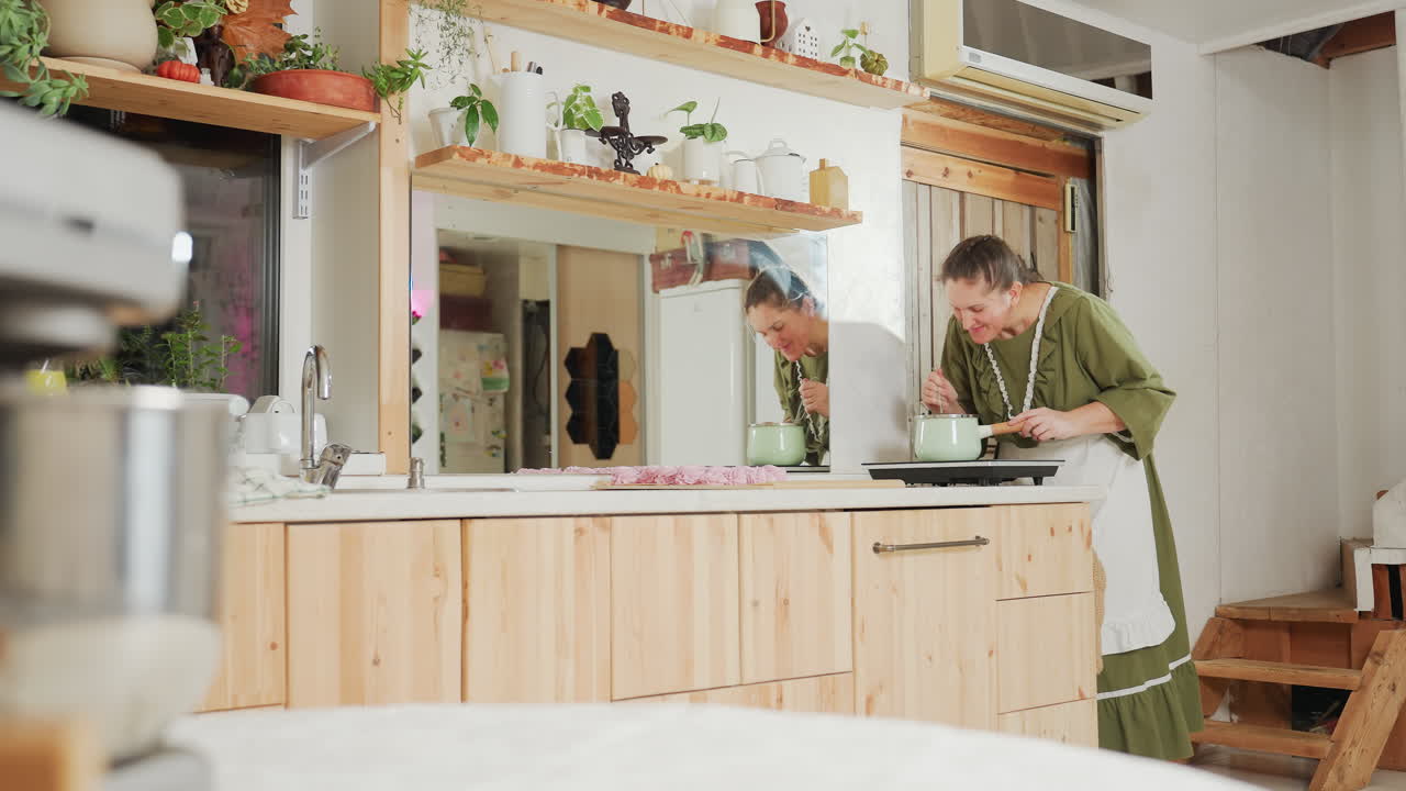 Housewife in green dress and apron leans over stove to smell aroma from pot while cooking dinner in cozy kitchen, with large wall mirror reflecting background plants, shelves, and kitchen decor