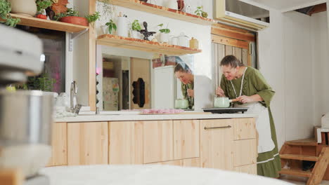 housewife in green dress and apron leans over stove to smell aroma from pot while cooking dinner in cozy kitchen, with large wall mirror reflecting background plants, shelves, and kitchen decor