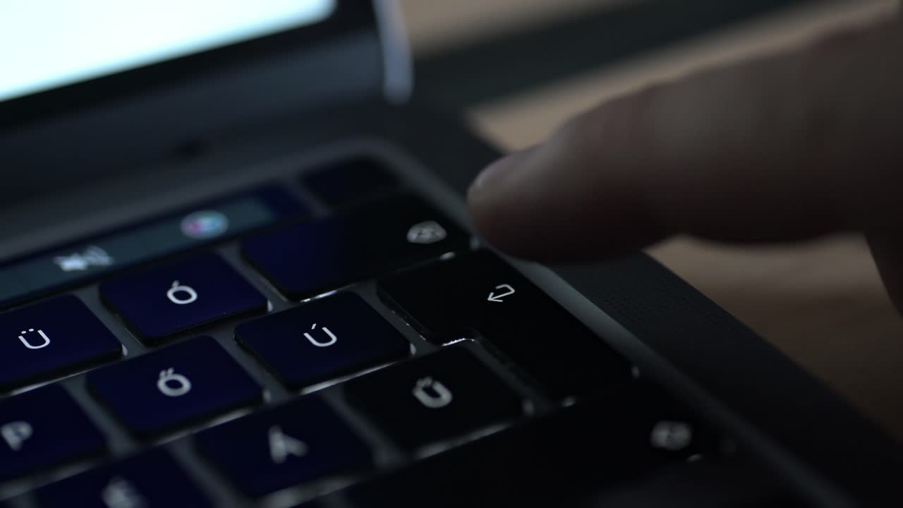 A close-up video showing a person's finger hovering above the enter key on a laptop keyboard with accented character keys. The scene is lit by soft, ambient light, creating a modern atmosphere