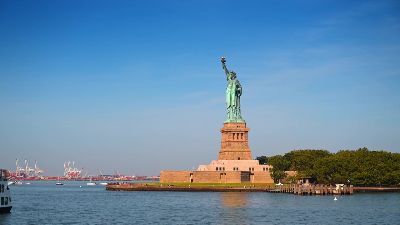 Patung Liberty di Pulau Liberty, Kota New York. Monumen tembaga itu menjulang di atas pepohonan dan air pelabuhan dengan derek-derek jauh di cakrawala.