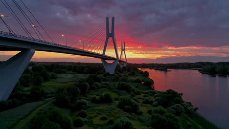 a stunning view of a modern cable-stayed bridge over a river at sunset or sunrise, with vibrant colors reflecting on the water and lush green banks