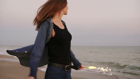 Young-beautiful-woman-running-by-the-sea-during-sunset-and-holding-burning-sparkling-candles-in-both-hands,-turning-around