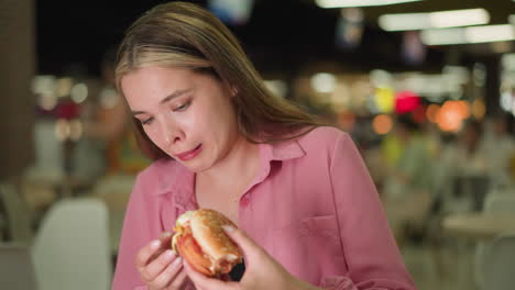 lady in pink dress examines burger with a displeased expression, showing signs of disgust before dropping it, people in the background are eating, with the view blurred and lights softly illuminating