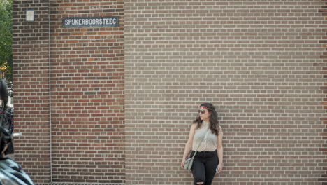 stylish woman with sunglasses leaning and posing in front of classical red brick stone wall in cozy town in netherlands in slowmo