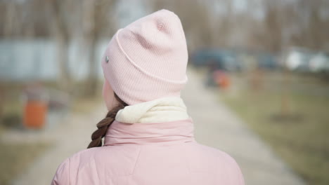 close-up rear view of young woman in light pink beanie, scarf, and winter jacket walking along paved path in quiet city park during cold season, surrounded by blurred trees and muted background