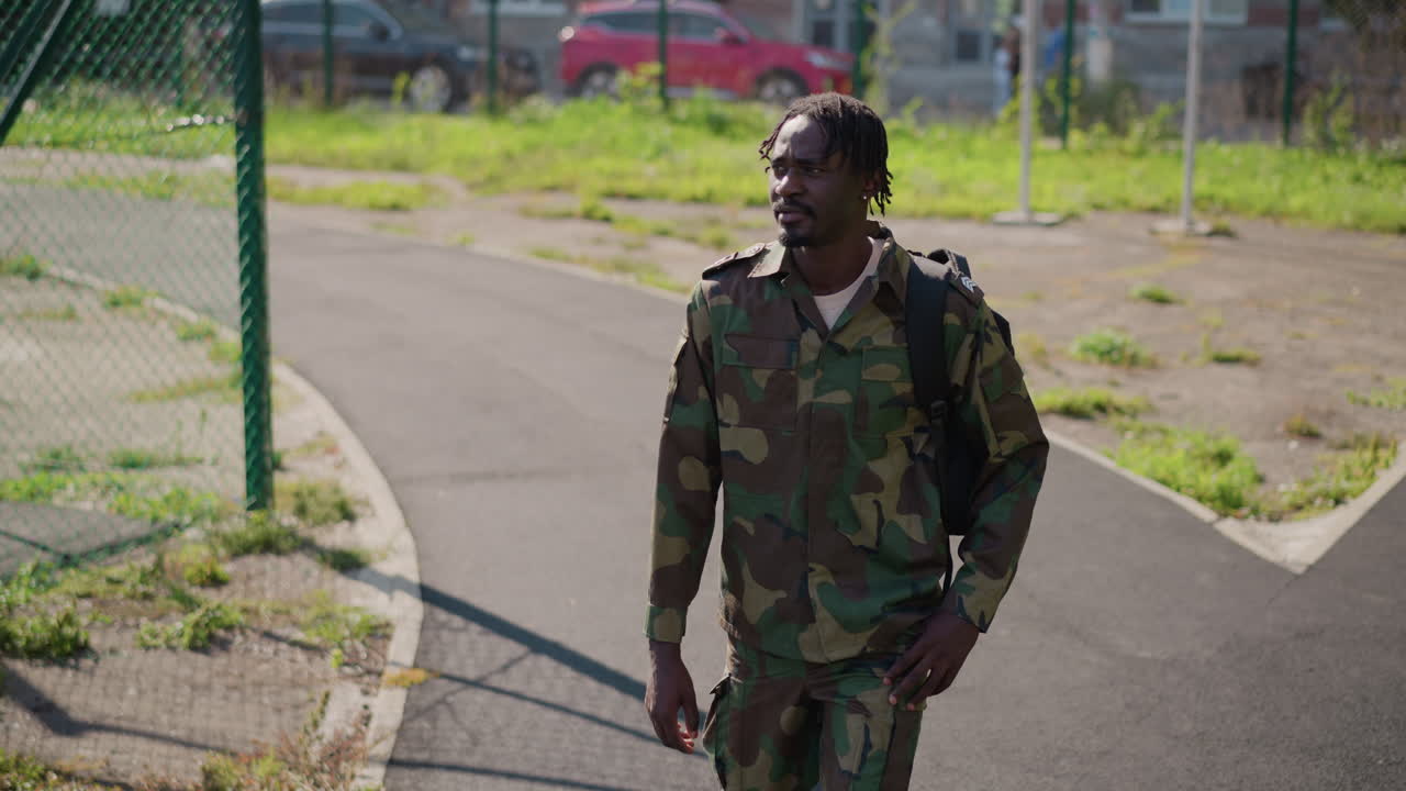 Soldier Walking Along Neighborhood Path, Camo Jacket And Backpack Near Chainlink Fence And Parked Cars, Sunlit Urban Park Edge, Cautious Gaze And Thoughtful Expression, Exploration Of Civilian Streets