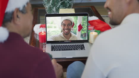 padre y hijo biraciales sonrientes usando una computadora portátil para una videollamada de navidad con un hombre en la pantalla