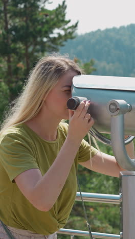 woman tourist in green t-shirt looks through large tower viewer standing on terrace at green highland resort on warm summer day slow motion