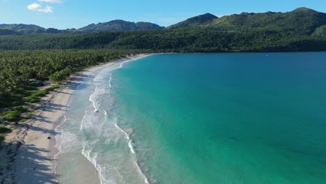Aerial backwards of clear Caribbean sea with tropical landscape and palm trees with exotic mountains in distance. Blue sky in summer season. Wide shot.