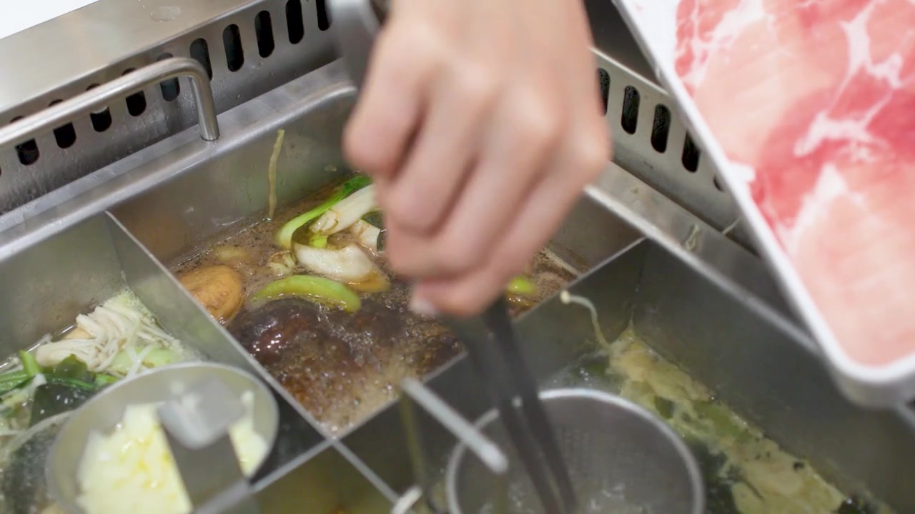 Närbild på en hand som använder tång för att tillaga rått fläskkött i en kokande restaurang-hot pot.