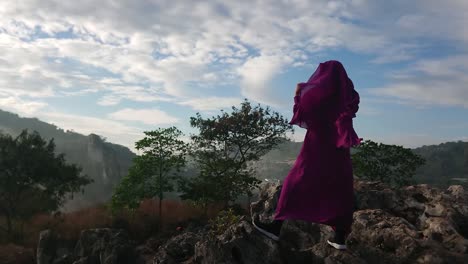 islamic woman wearing purple burqa stands on rocky summit looking at landscape